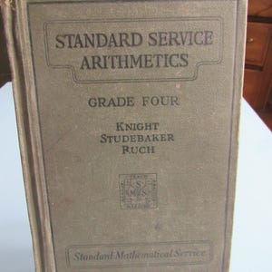 May include: A vintage hardcover book titled "Standard Service Arithmetics, Grade Four" with the authors Knight, Studebaker, and Ruch. The cover is a muted olive green with black lettering and a small emblem. The book is a relic of educational materials.