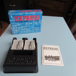 May include: A blue and white box with the text "Express The Railroad Card Game" on it. The box is next to a black card holder with two cards inside. The card holder has the text "Express The Railroad Card Game" on it. There is also a white instruction booklet with the text "Express The Railroad Card Game" on it.