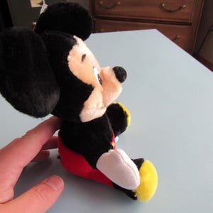 May include: A plush Mickey Mouse toy, featuring black ears, a black head, and a tan face. The toy is wearing red shorts, white gloves, and yellow shoes. The toy is sitting on a light blue surface.