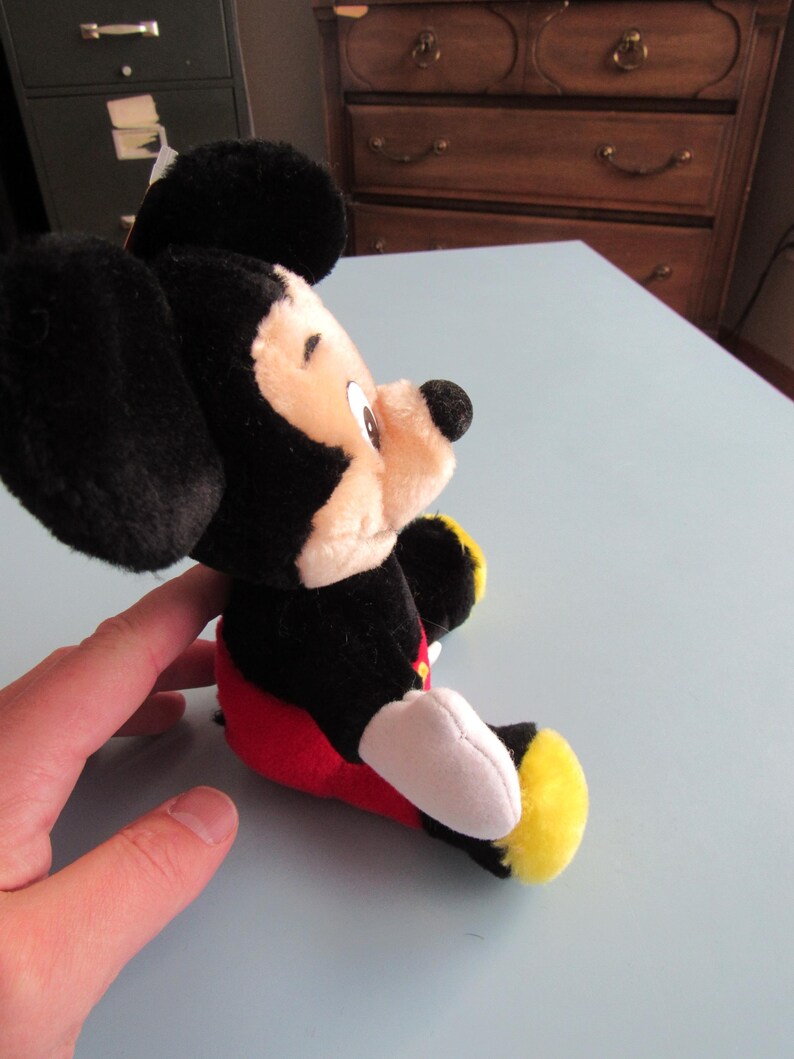 May include: A plush Mickey Mouse toy, featuring black ears, a black head, and a tan face. The toy is wearing red shorts, white gloves, and yellow shoes. The toy is sitting on a light blue surface.