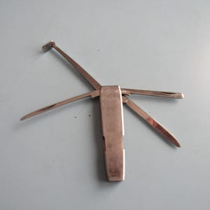 May include: A silver-toned multi-tool featuring a central rectangular handle with four distinct metal implements extended outward. The tools include a flat-ended scraper, a pointed blade, a thin probe, and a narrow spatula, all made of polished metal.
