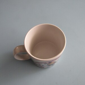 May include: A ceramic mug with a light-colored interior and a handle on the side. The exterior features a decorative band with a graphic illustration of a character, showing a circular opening at the top and a smooth, rounded base structure.