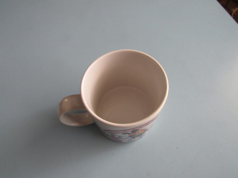 May include: A ceramic mug with a light-colored interior and a handle on the side. The exterior features a decorative band with a graphic illustration of a character, showing a circular opening at the top and a smooth, rounded base structure.