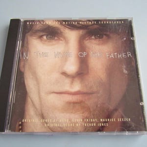 May include: A CD case featuring a close-up of a man's face. The title "IN THE NAME OF THE FATHER" is printed in white across the top. The case also includes text indicating it is a soundtrack with original songs and score.