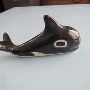 May include: A ceramic orca figurine, predominantly black with white accents on the belly and eye. The orca has a glossy finish and is posed in a swimming position. The figurine is a decorative item.