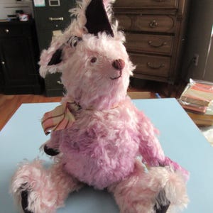 May include: A pink plush toy dog with a dark brown hat and paws. The dog has a light pink body and a striped scarf around its neck. The toy is sitting on a light blue surface.