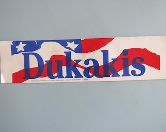 May include: Rectangular banner with the name "Dukakis" in blue, set against a backdrop of a stylized American flag in red, white, and blue. The banner includes text at the bottom, likely related to the campaign.