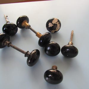 May include: A collection of vintage black door knobs with metal stems and brass accents. Some knobs are oval-shaped, while others are round. One knob shows signs of wear with peeling paint, revealing a lighter color underneath.
