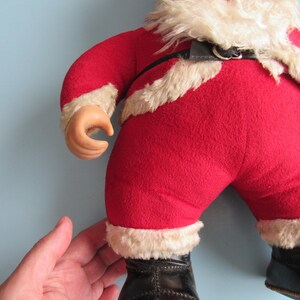 May include: A vintage Santa Claus doll with a red suit trimmed with white faux fur. The doll has a black belt, black boots, and a white beard. The doll's face is made of a hard material with painted features.