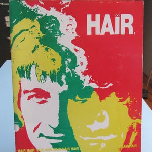 May include: A vintage book cover with the word "HAIR" in white block letters on a red background. The cover features a stylized illustration of two faces with green, yellow, and white accents. The words "HAIR" are repeated at the bottom.