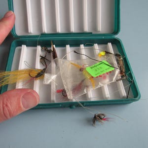 May include: An open, teal-colored fishing tackle box filled with various colorful fishing flies. The flies have feathers, thread, and hooks. A green tag with the word "Walgreens" is visible. The box is on a light blue surface.