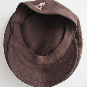 May include: A brown Kangol cap, seen from the inside, revealing the inner lining and the brim. The Kangol logo, a kangaroo, is displayed on the front. The cap is made of a textured fabric.