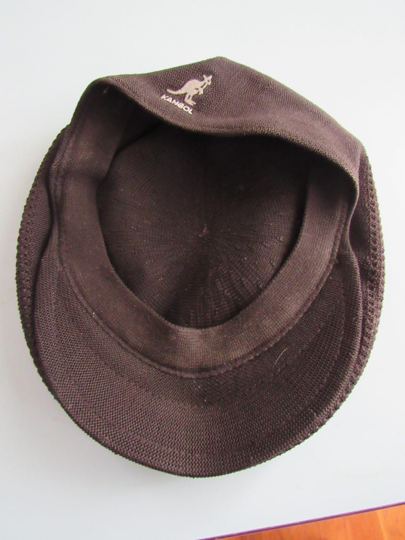 May include: A brown Kangol cap, seen from the inside, revealing the inner lining and the brim. The Kangol logo, a kangaroo, is displayed on the front. The cap is made of a textured fabric.