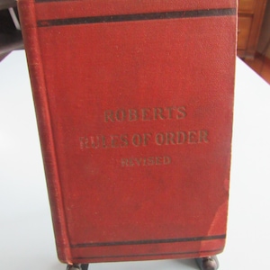 May include: A vintage red book titled "Roberts Rules of Order Revised." The book has black horizontal stripes and gold lettering. It is propped up on small, dark brown feet.