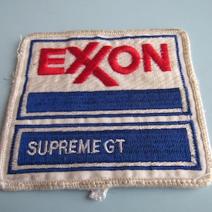 May include: Embroidered patch featuring the Exxon logo in red letters over a white background. Below the logo is a blue rectangle with the words "SUPREME GT" in white. The patch has a white border.