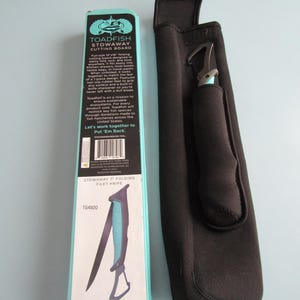 May include: A black Stowaway 7" folding filet knife with a blue handle and a black sheath. The packaging is a light blue box with the text "TOADFISH STOWAWAY CUTTING BOARD" and "STOWAWAY 7 FOLDING FILET KNIFE".