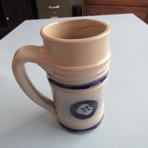 May include: A beige ceramic mug with a handle. The mug features a dark blue emblem and blue stripes. The mug is cylindrical with a wider top and a textured surface. The mug is likely used for drinking beverages.