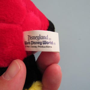 May include: A close-up of a plush toy with red, black, and yellow coloring. The tag reads "Disneyland Walt Disney World &copy; Walt Disney Productions."