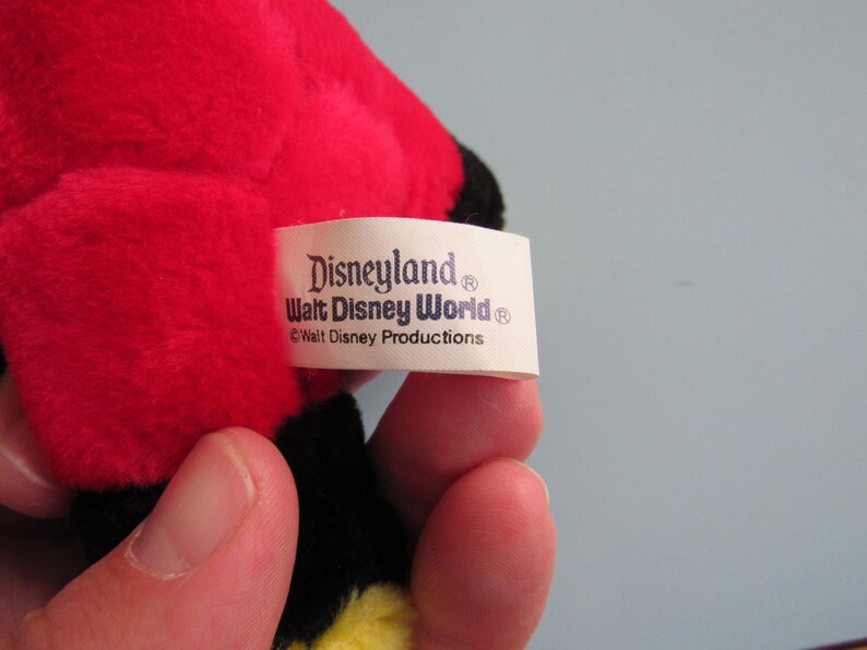 May include: A close-up of a plush toy with red, black, and yellow coloring. The tag reads "Disneyland Walt Disney World &copy; Walt Disney Productions."