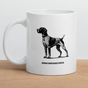 Puede incluir: Taza de cerámica blanca con una ilustración en blanco y negro de un Braco Alemán. El perro se muestra de pie, mostrando sus marcas distintivas. El texto "GERMAN SHORTHAIRED POINTER" está impreso debajo.