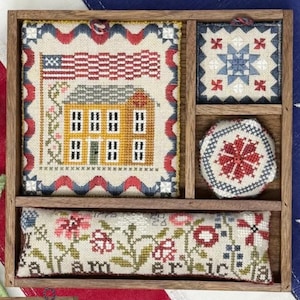 May include: A wooden framed display featuring four cross-stitch pieces with patriotic themes. The pieces include an American flag, a house, a star pattern, and a floral design with the words "I am America".