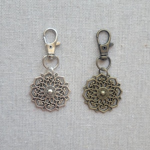 May include: Two decorative keychains with ornate, floral-inspired pendants. One keychain is silver-toned, the other is bronze. Each has a lobster clasp for easy attachment. The pendants feature a central starburst design.