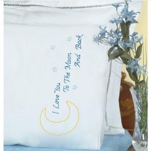 May include: White pillowcase with embroidered text "I Love You To The Moon And Back", a yellow crescent moon, and blue stars. A vase of blue flowers is in the background. The pillowcase has a decorative stitched border.
