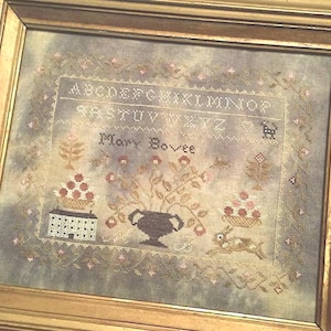 May include: Framed cross-stitch sampler with a gold-colored wooden frame. The sampler features the alphabet, the name "Mary Bovee", and various floral and animal motifs in muted colors. The design includes a rabbit, flowers, and a house.