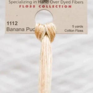 May include: Close-up of a skein of cotton floss in a light beige color, labeled "Banana Puc." The label also indicates the floss is 5 yards long and part of the "FLOSS COLLECTION." The text "Specializing in Hand Over Dyed Fibers" is also visible.