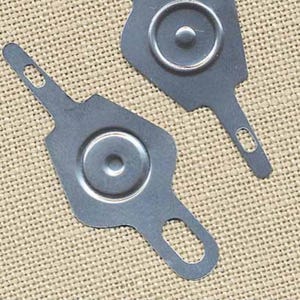 May include: Two silver-colored metal tools with a circular design and a handle with a hole. The tools are on a tan, textured surface. The tools have a key-like shape.
