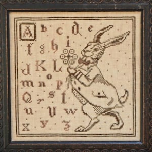 May include: A framed cross-stitch sampler featuring a rabbit holding flowers, surrounded by an alphabet. The rabbit is wearing a suit. The sampler is in shades of brown and beige, with a dark brown frame. The style is vintage and folk art.