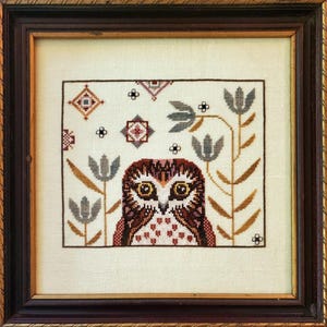 May include: Framed cross-stitch artwork featuring a detailed owl with large yellow eyes, surrounded by stylized flowers and geometric patterns. The owl is rendered in shades of brown, red, and white, set against a cream-colored background. The frame is dark brown.
