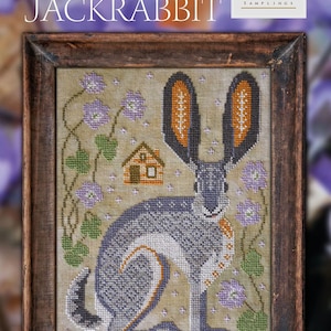 May include: A framed cross-stitch design of a gray jackrabbit with large ears, sitting in a field of purple flowers and green leaves. The design is titled "The Jackrabbit" and is part of a series called "A Year in the Woods #3".