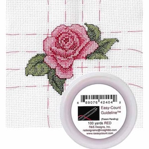 May include: Cross-stitch project featuring a pink rose with green leaves on white fabric. A spool of red thread is in the foreground. The spool label reads "Easy-Count Guideline" and "100 yards RED".