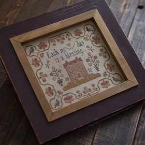 May include: A framed cross-stitch sampler with a beige background and a pink house with the text "Each new day is a blessing".