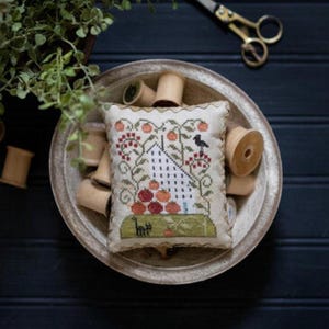 May include: A rectangular cream-colored pincushion featuring a cross-stitch design of a white house, red tomatoes, green vines, a black bird, and a black cat. The pincushion is centered in a shallow bowl filled with wooden spools of thread.
