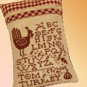 May include: A rectangular pillow with a tan background and a red and brown checkered top. The pillow features a cross-stitched turkey design, alphabet, and the words "TOM TURKEY CT 2018".