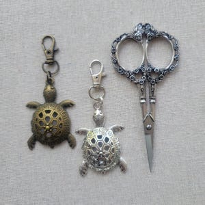 May include: Two turtle-shaped keychains, one bronze-toned and one silver-toned, with lobster claw clasps. A pair of ornate, silver-toned scissors with floral detailing is also present. The items are displayed on a neutral fabric background.