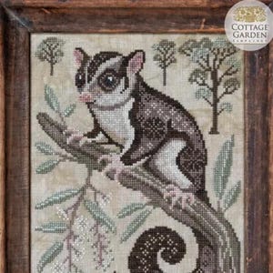 May include: Cross-stitch artwork of a sugar glider perched on a branch, surrounded by leaves and small trees. The sugar glider has a white and brown body with large eyes. The artwork is framed in a dark wooden frame.