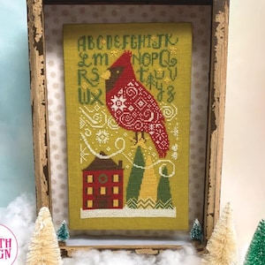 May include: Cross-stitch artwork of a red cardinal with a detailed pattern, perched atop stylized green and yellow trees, and a red house. The background is a light green color with embroidered letters and snowflakes. The artwork is framed in a rustic wooden frame.