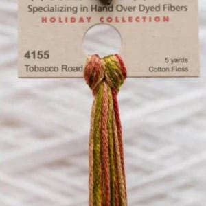 May include: A skein of cotton floss in shades of red, green, and brown, with a tag that reads "HOLIDAY COLLECTION." The tag also indicates the color number "4155 Tobacco Road" and 5 yards of floss.