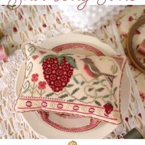May include: A white pillow with a cross-stitch design of a strawberry, leaves, and a bird. The pillow is on a white plate with a pink and red border. The text "Strawberry Faire" is at the top of the image. The text "october house fiber arts" is at the bottom of the image.