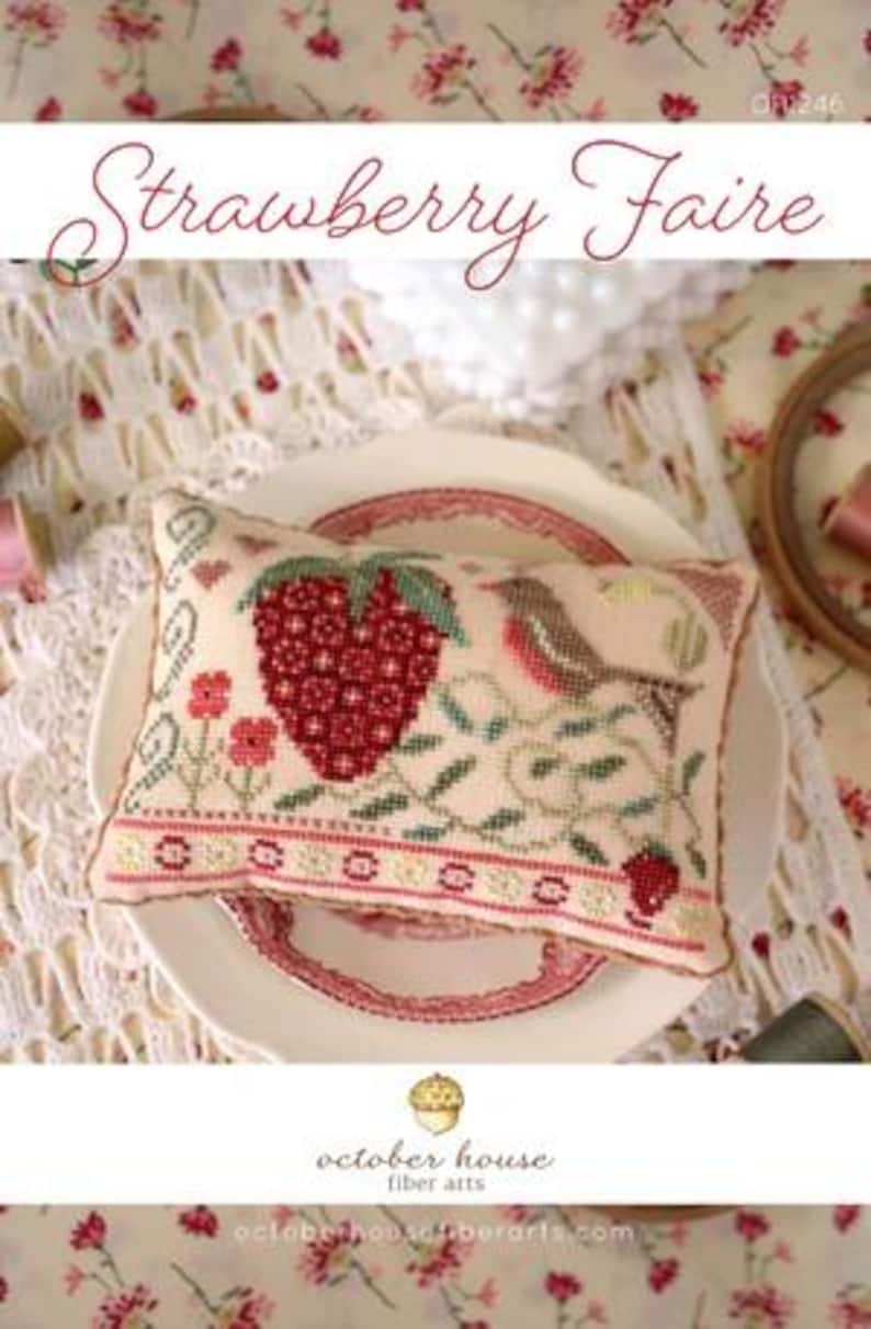May include: A white pillow with a cross-stitch design of a strawberry, leaves, and a bird. The pillow is on a white plate with a pink and red border. The text "Strawberry Faire" is at the top of the image. The text "october house fiber arts" is at the bottom of the image.