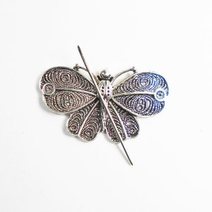 May include: A silver-toned butterfly brooch with intricate filigree detailing. The wings feature a delicate openwork design, and the body has a textured appearance. The brooch has a pin on the back for secure fastening. The butterfly is a classic jewelry motif.