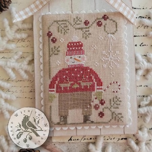 May include: Cross-stitch artwork of a snowman wearing a red sweater and hat, holding a candy cane. The artwork is framed with a scalloped edge and a beige ribbon. A circular emblem with a bird is in the lower left corner.