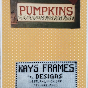 May include: A handmade cross-stitch design with the word "PUMPKINS" in red letters, framed in a dark green border. Below, a second cross-stitch reads "KAYS FRAMES AND DESIGNS, WESTLAND, MICHIGAN". The background is yellow with white polka dots.