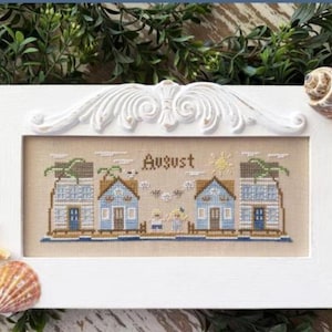 May include: A framed cross-stitch piece with a beach theme, featuring blue houses and the word "August." The white frame has decorative accents and is adorned with seashells and a starfish.