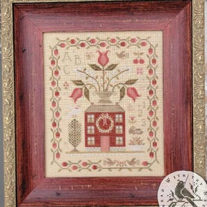 May include: Framed cross-stitch art featuring a house with a wreath, a vase of flowers, and a border of berries. The artwork is in a wooden frame with a gold-colored inner edge. The color palette includes red, beige, and green.