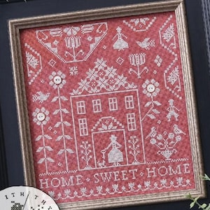 May include: Framed cross-stitch art with a red background and white stitching. The design features a house, flowers, birds, and the text "HOME SWEET HOME." The artwork is in a black frame with a gold inner border, showcasing a traditional craft.
