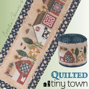 May include: A cross-stitch design featuring a "Quilted Tiny Town" theme. The design includes houses, trees, and floral elements in a rustic style. A matching round pincushion is also shown. The color palette includes beige, red, blue, and green.